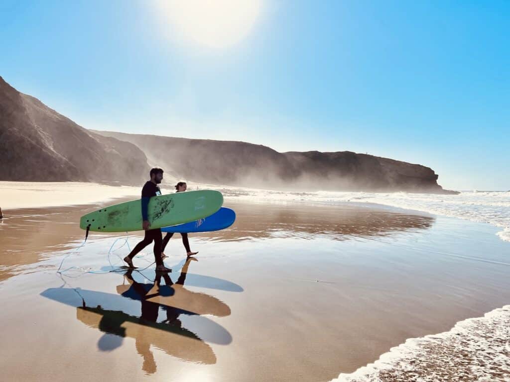 Surfisti con tavole sulla spiaggia Due surfisti trasportano tavole da surf verdi e blu lungo la spiaggia quando il tempo è soleggiato.